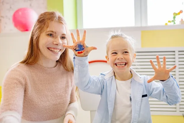 happy student showing hands covered in paint next to smiling teacher